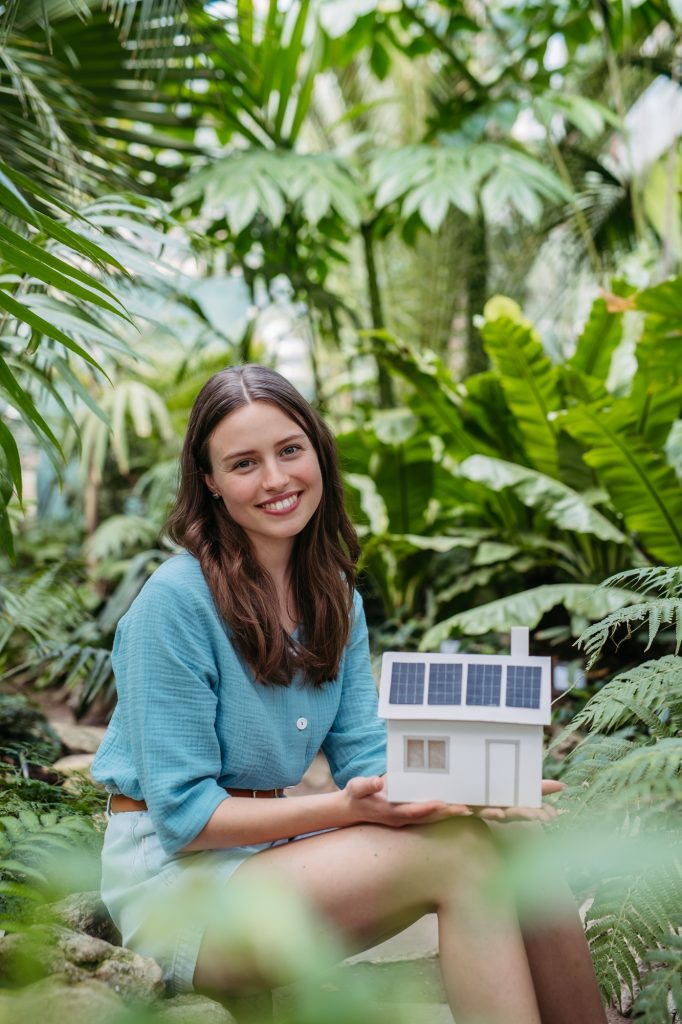 Young woman in jungle holding paper model of house with solar panels, concept of renewable energy
